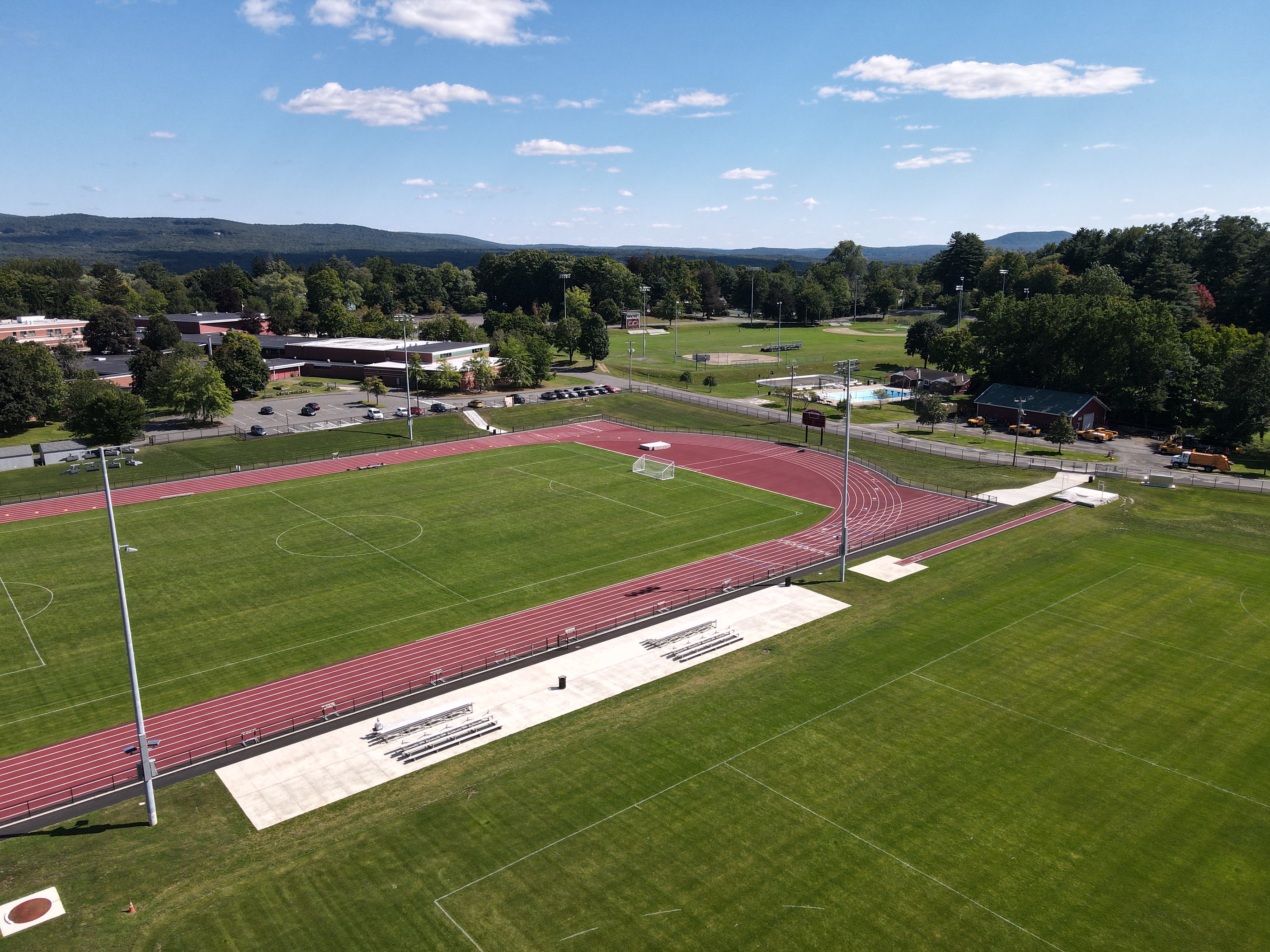 Aerial view of a sports field featuring a soccer pitch and a running track, surrounded by green grass and trees, with a school building and parking lot in the background.