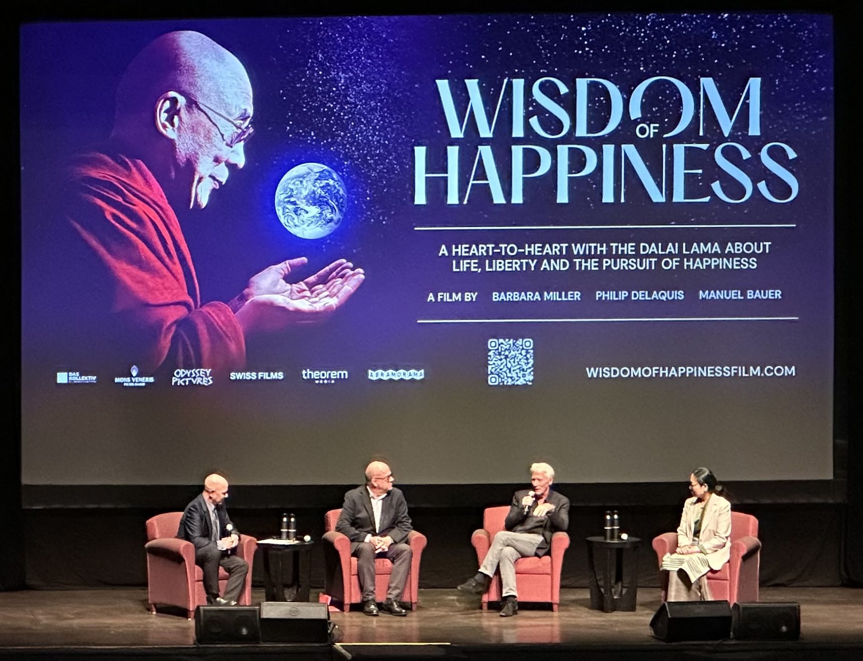 Promotional image for the film 'Wisdom of Happiness' featuring a graphic of the Dalai Lama, along with four speakers seated on stage discussing the themes of life, liberty, and the pursuit of happiness.