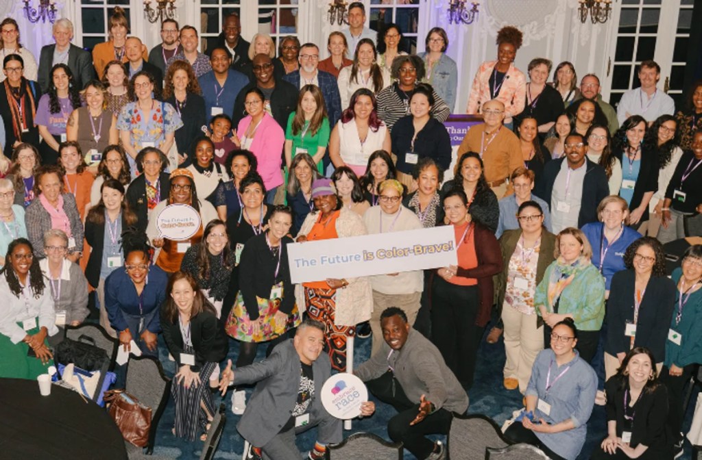 Group photo of diverse attendees at an event, holding a sign that reads 'The Future is Color-Brave!'