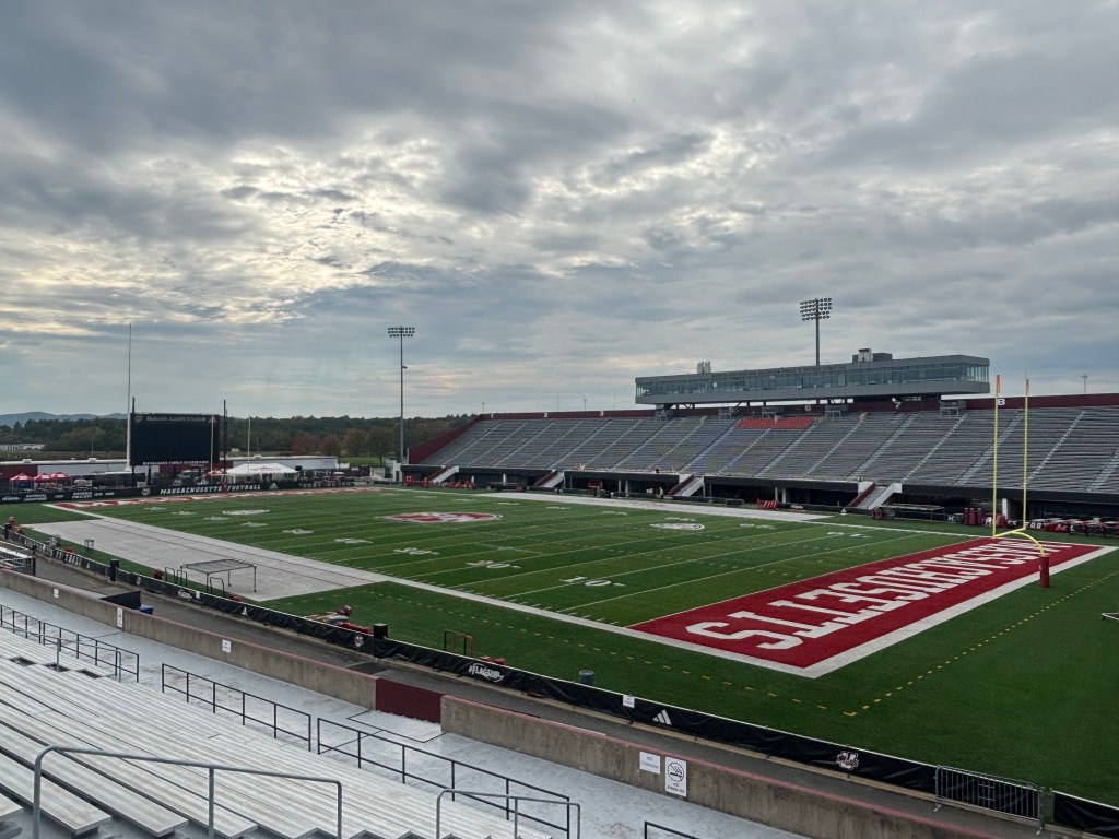 A view of a football stadium featuring green turf marked with yard lines and a prominent logo, empty grandstands on the left, and a large scoreboard in the background under a cloudy sky.