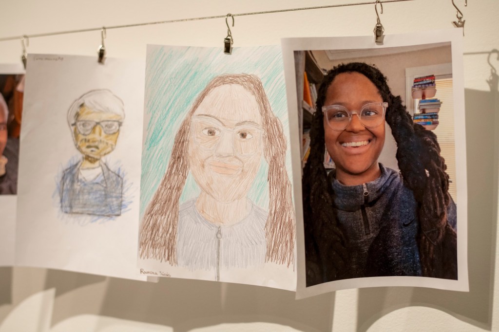 An art display featuring two portraits: a hand-drawn sketch of a person with glasses and another hand-drawn portrait of a smiling person with long hair, alongside a photograph of the smiling person.