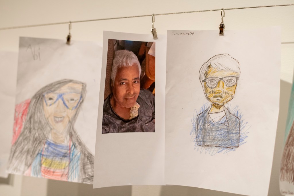 A display of three portraits: a child's drawing of a person with glasses and long hair, a photograph of a man with gray hair, and another child's drawing of a man with glasses and short hair, all hanging from a string.