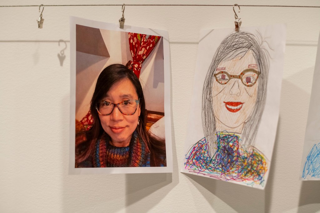 A photograph of a woman with long dark hair and glasses, smiling, next to a colorful childlike drawing of the same woman with glasses and a wide smile, hanging on a wall.