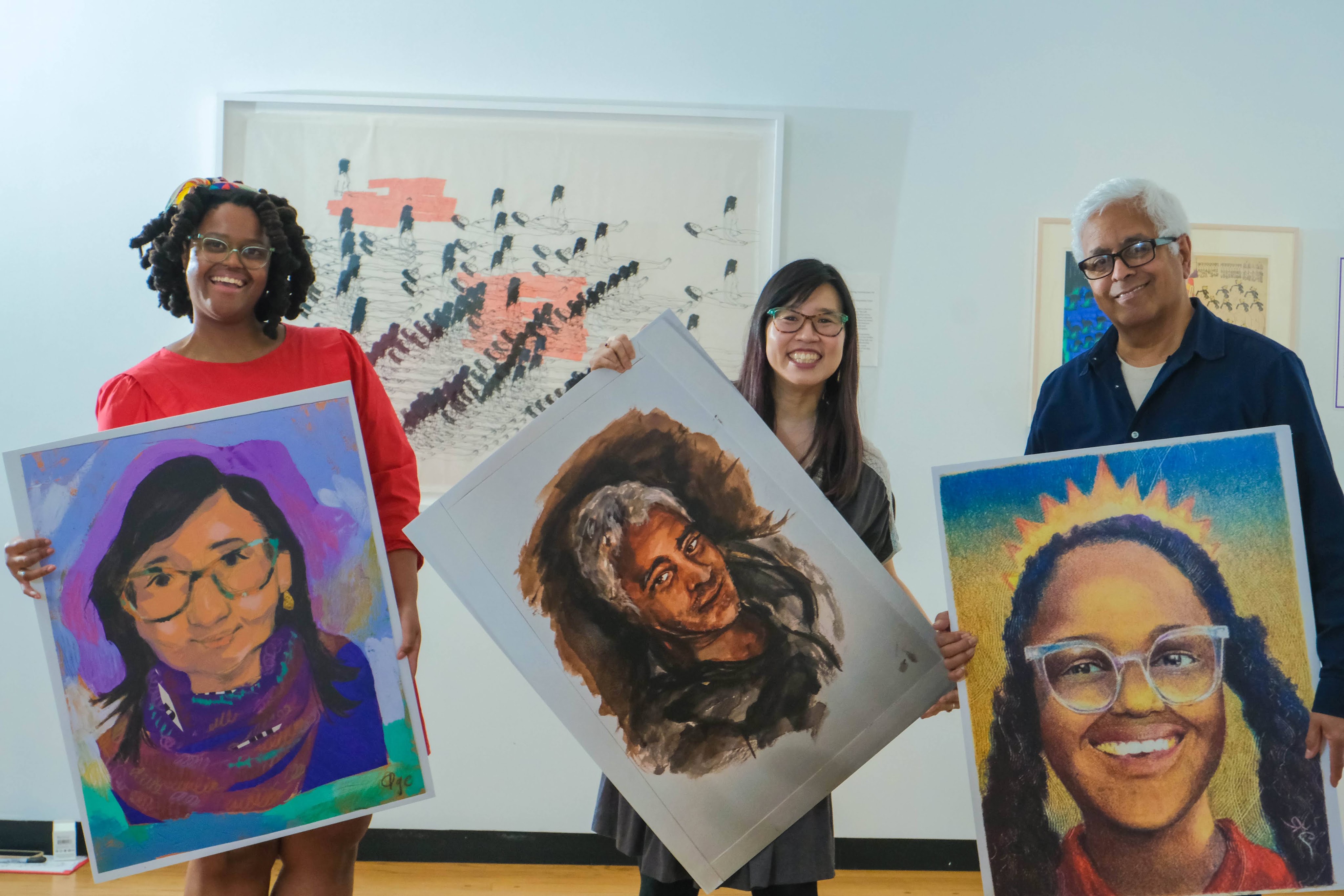 Three individuals hold colorful portrait paintings indoors, showcasing diverse artistic styles. The background features abstract artwork.